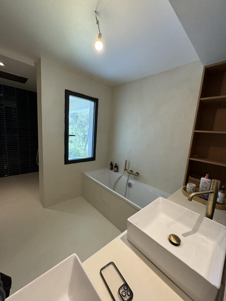 Salle de bain moderne crème et bois avec grande douche noire, baignoire sous fenêtre, deux lavabos blancs et robinetterie laiton.