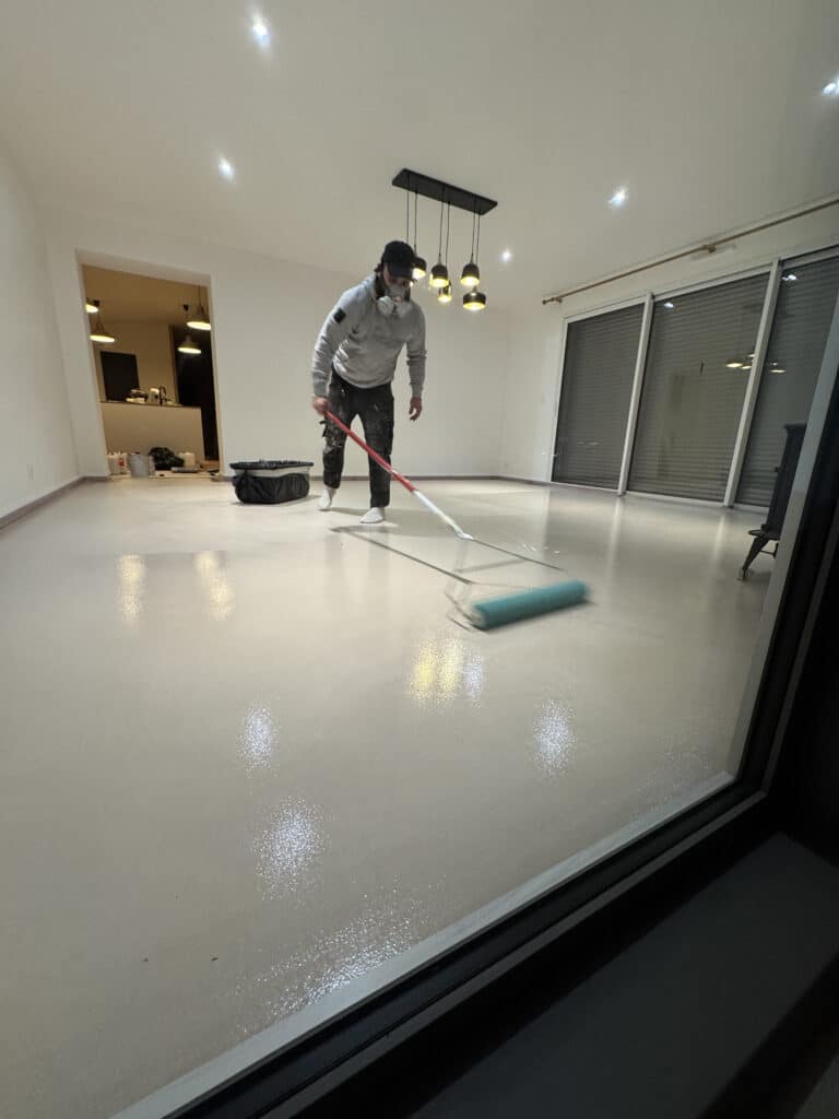 Homme masqué appliquant un revêtement de sol clair brillant avec un rouleau dans une pièce vide et lumineuse.