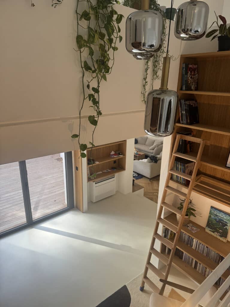 Vue plongeante sur un intérieur moderne avec plantes suspendues, luminaires design, grande bibliothèque et porte-fenêtre donnant sur une terrasse.