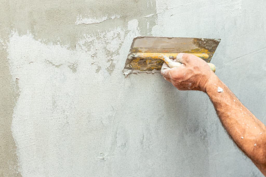 Main d'artisan appliquant de l'enduit frais sur un mur texturé avec une truelle, dans un processus de rénovation.