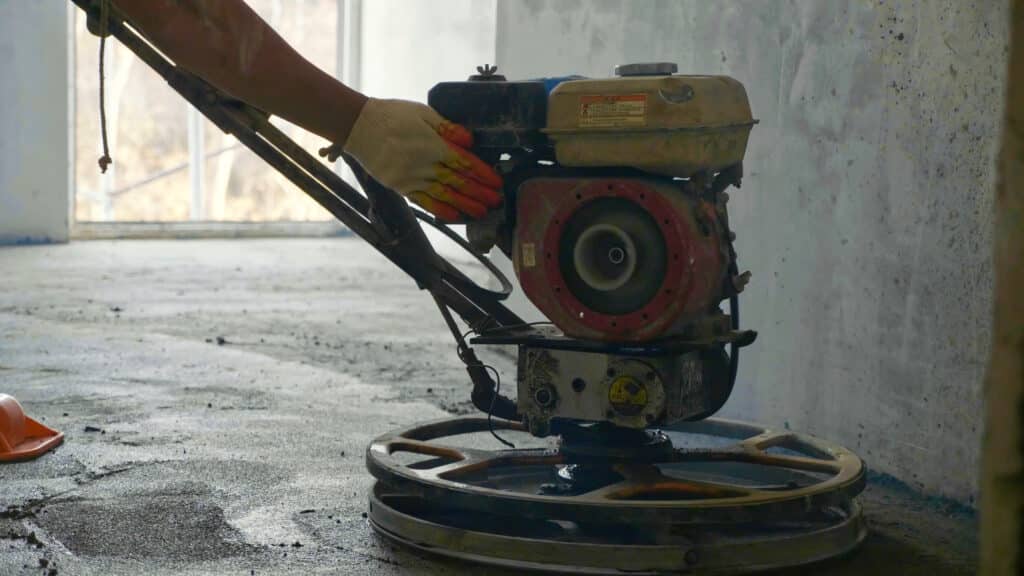 Ouvrier ganté utilisant une polisseuse à béton thermique sur sol en ciment. Réservoir d'essence visible avec étiquette d'avertissement.