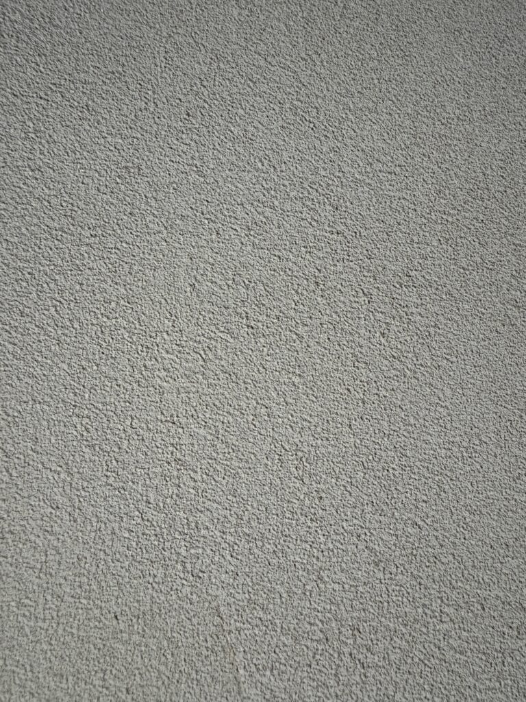 Surface de mur grise à texture rugueuse. Crépi granuleux avec ombres légères, créant un fond abstrait et moderne.
