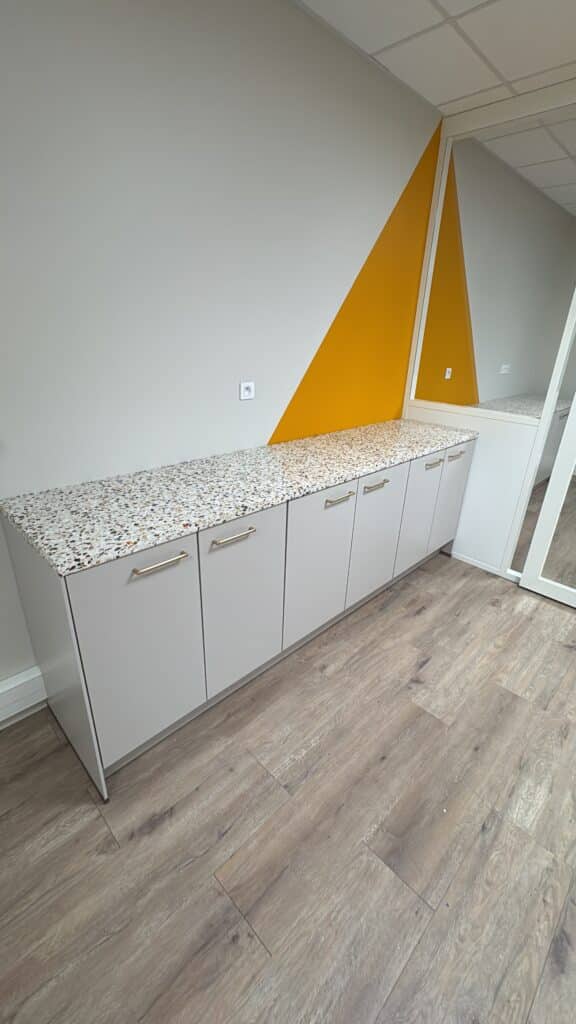 Rangement bureau moderne : meuble gris, terrazzo et mur jaune Meuble bas gris moderne à six portes, surmonté d'un comptoir en terrazzo moucheté. Mur géométrique gris et jaune vif contrastant.