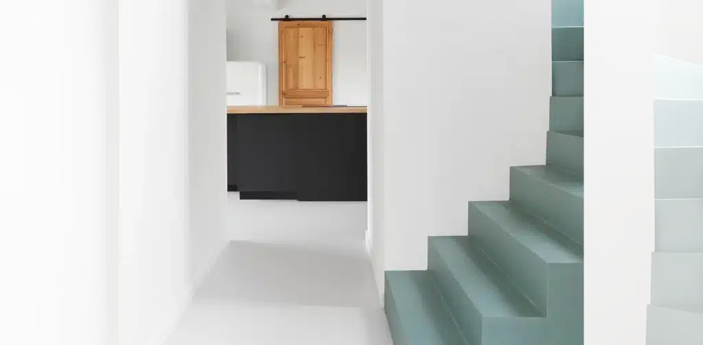 Escalier moderne bleu-vert minimaliste en contraste avec un couloir blanc menant à une cuisine noire et bois avec porte coulissante.