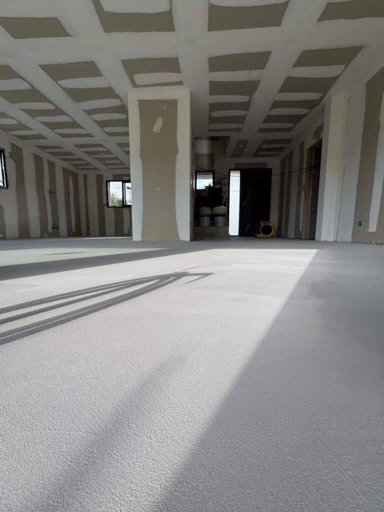 Sol d'un chantier neuf avec revêtement gris texturé, ombres projetées, murs et plafond en plaques de plâtre non finies.