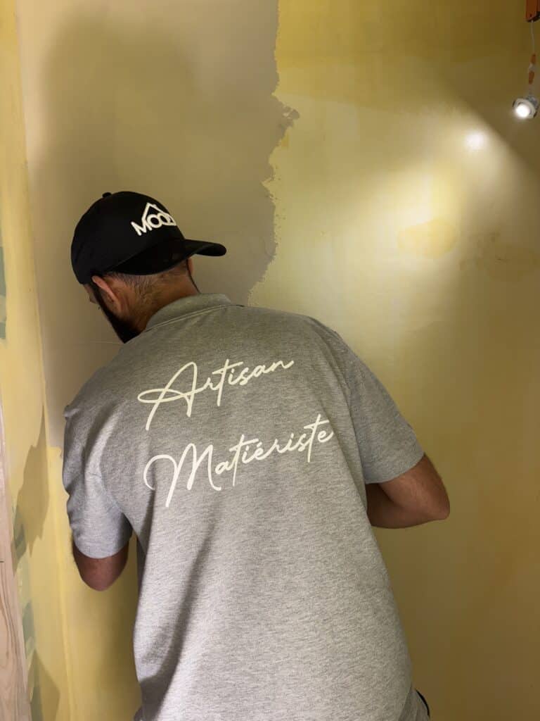 Artisan Matieriste au travail sur mur jaune Artisan Matieriste en t-shirt gris et casquette noire appliquant un enduit mural jaune, travail en cours.