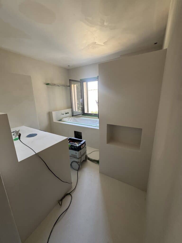 Chambre de bain en construction, murs plâtrés de couleur crème, baignoire encastrée, fenêtre ouverte, aspirateur Festool au sol.