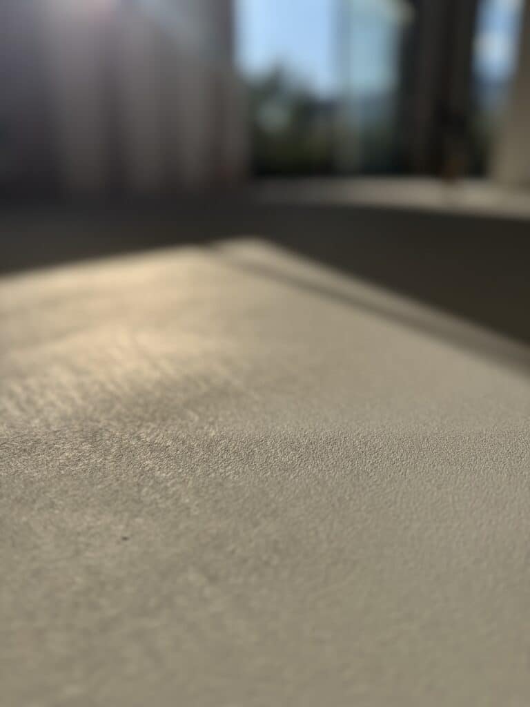 Gros plan sur une surface texturée beige-gris, éclairée par un reflet de lumière rasante venant d'une fenêtre floue en arrière-plan.