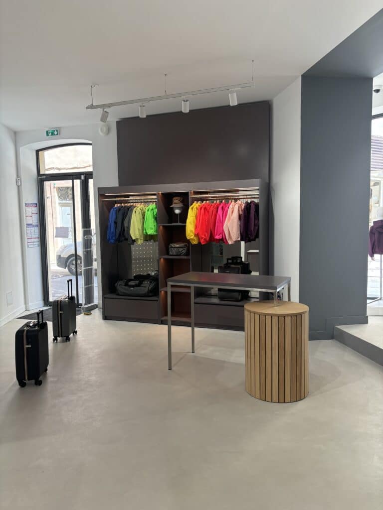 Boutique K-Way au design gris et bois. Présentoir de vestes colorées (rouge, jaune, vert), deux bagages à roulettes, et consigne de sécurité (Manipulation des extincteurs).