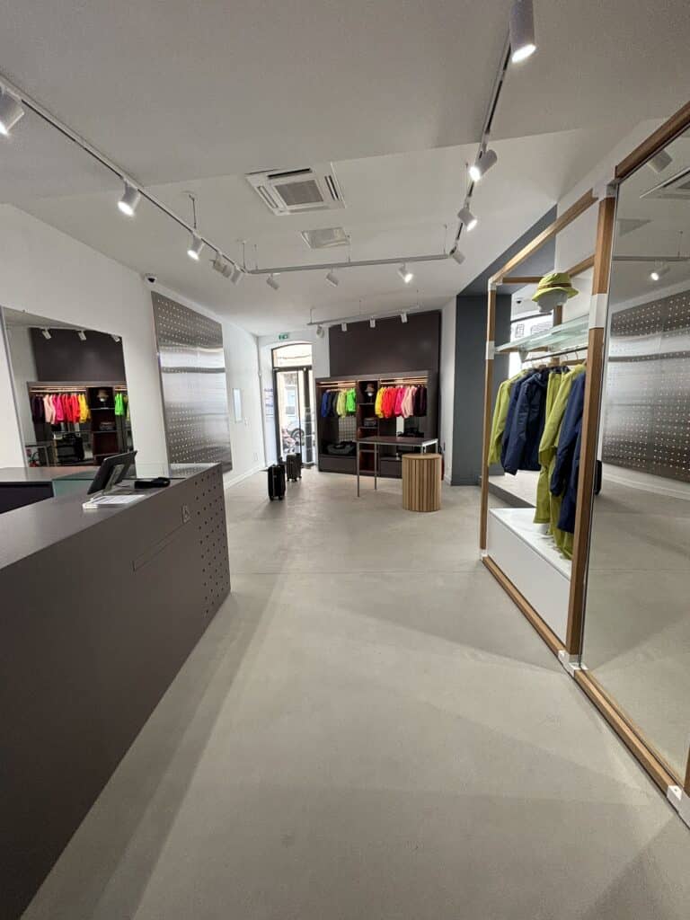Intérieur boutique design moderne. Comptoir gris percé, sol béton ciré. Présentoirs avec vestes colorées fluo sous éclairage sur rail.