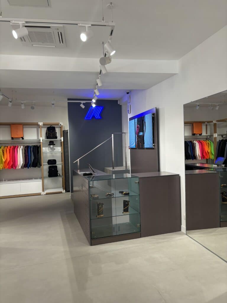 Boutique K-Way moderne. Vêtements imperméables colorés, sacs, et accessoires exposés. Logo néon bleu et comptoir en verre minimaliste.
