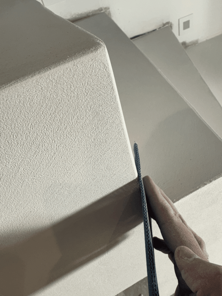 Main ponçant l'angle d'un mur à l'enduit texturé blanc pour des finitions de rénovation soignées.