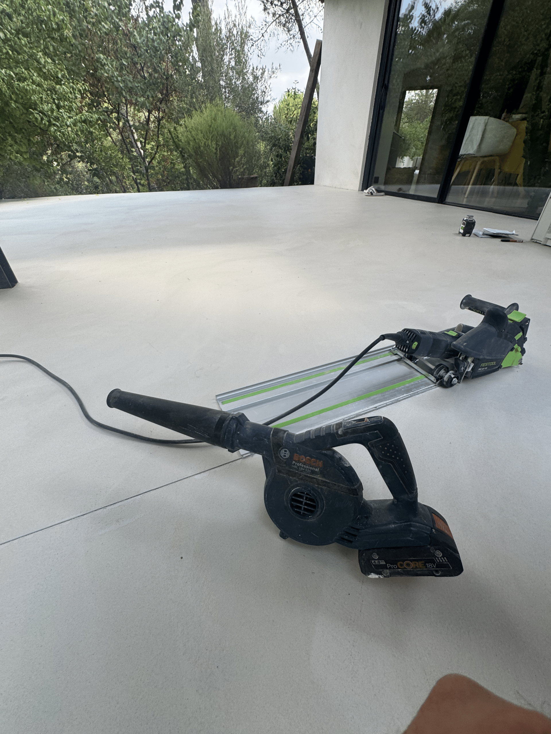 Outils Bosch et Festool sur Terrasse : Chantier Extérieur Souffleur Bosch GBL 18V-120 et scie Festool sur rail sur une terrasse en béton en plein air.
